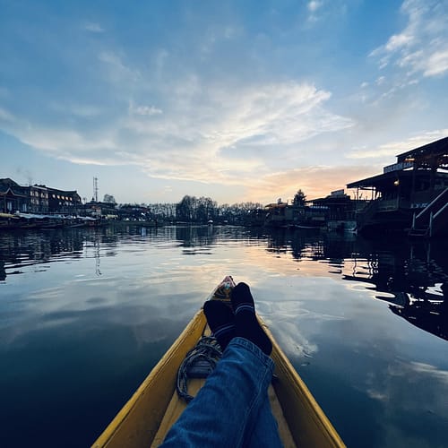 Person wearing a blue jeans reclining in a yellow boat on calm water at dusk, with silhouette of riverside houses against a cloudy sky, Kashmir Dale Lake.