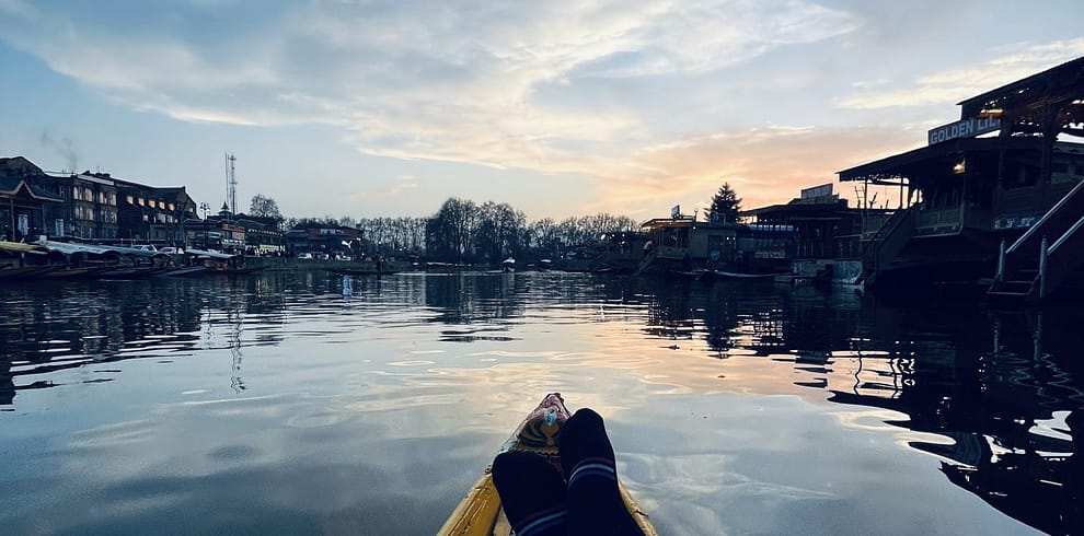 Person wearing a blue jeans reclining in a yellow boat on calm water at dusk, with silhouette of riverside houses against a cloudy sky, Kashmir Dale Lake.